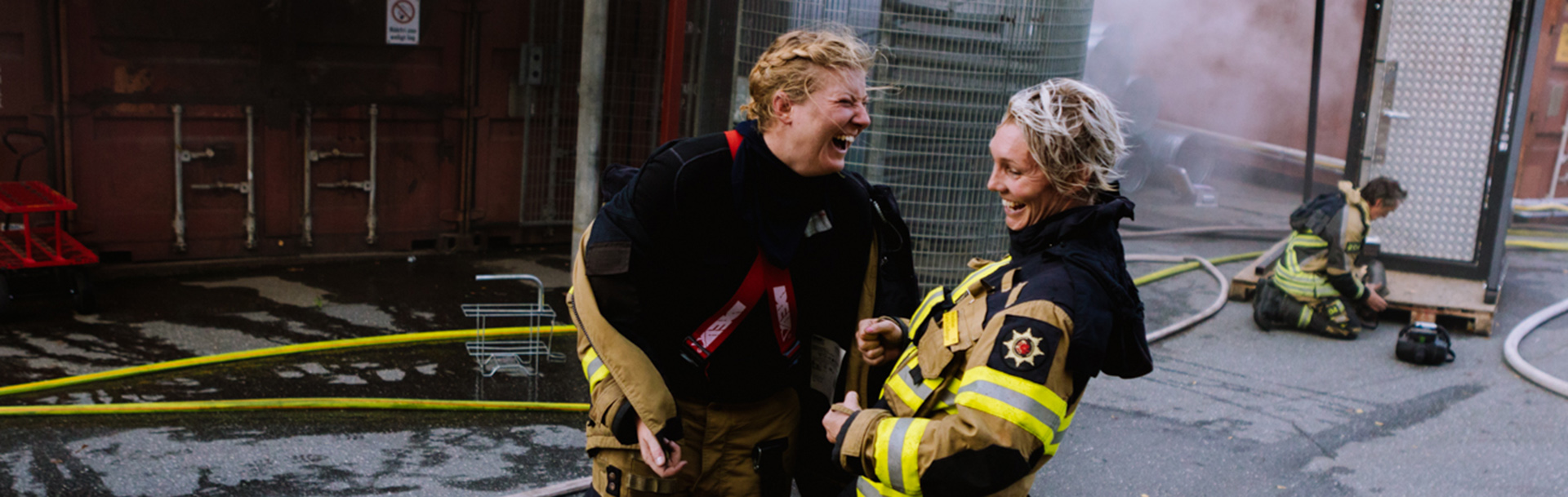 Två brandmän på övning som skrattar tillsammans
            
